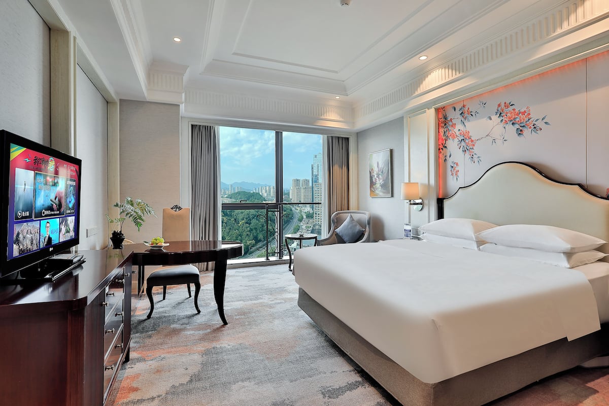 A hotel room with a large bed, a desk with a TV, a chair, a small table, and a view of the city.
