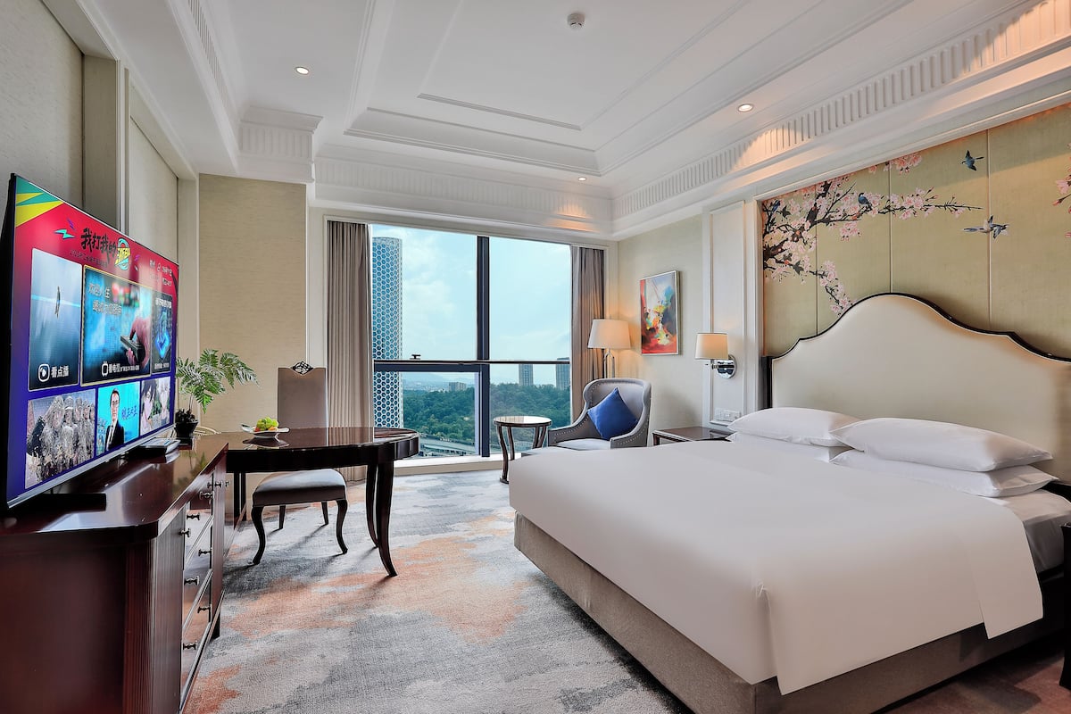 A hotel room with a large bed, a desk with a TV, a chair, a small table, and a view of the city.