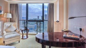 A hotel room with a large window, a piano, a chair, a small table with a plant, and a phone on the wall.