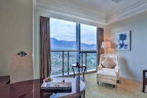 A hotel room with a large window, a chair, a table with a tray, and a lamp.