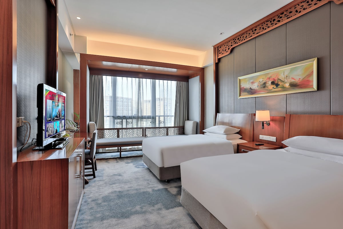 A hotel room with two beds, a TV, a desk, and a large window.