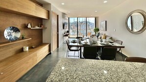 A modern dining area with a granite countertop, wooden shelving, and a large window offering a city view. - Boca de cielo Apartments (Mazatlán)