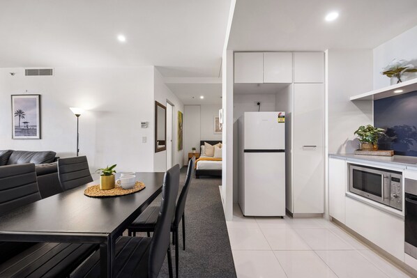 Una cocina moderna con armarios blancos, microondas integrado y refrigerador. Hay un comedor con mesa y sillas negras, y una pequeña planta en la mesa.