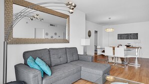 Ein modernes Wohnzimmer mit einem grauen Ecksofa, einem großen Spiegel und einem Essbereich mit einem Glastisch und weißen Stühlen.