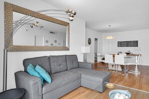 Moderni olohuone, jossa on harmaa kulmasohva, suuri peili sekä ruokailutila, jossa on lasipöytä ja valkoiset tuolit.