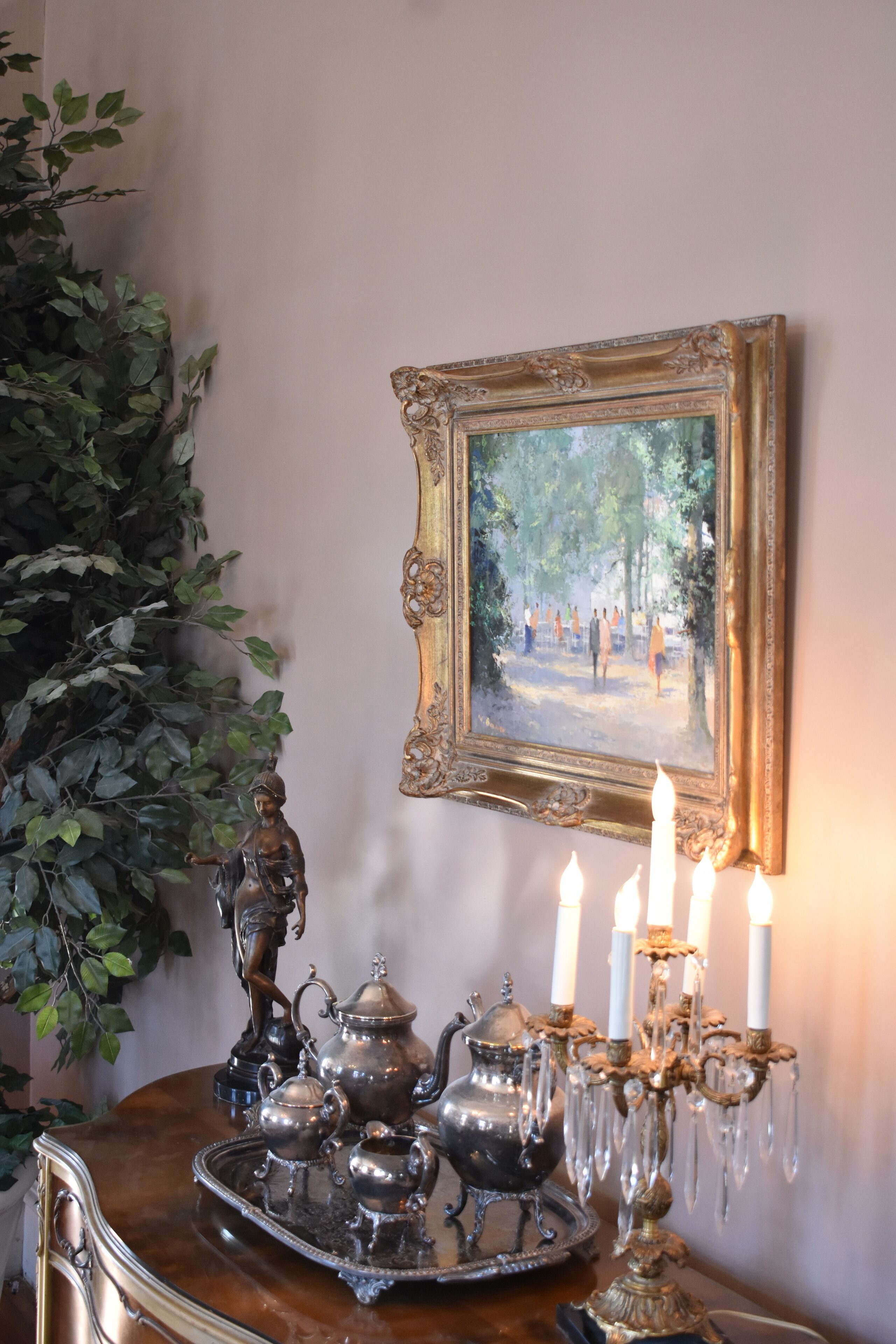 Una mesa con una bandeja de vajillas de plata, un candelabro con velas y un cuadro enmarcado de un parque.