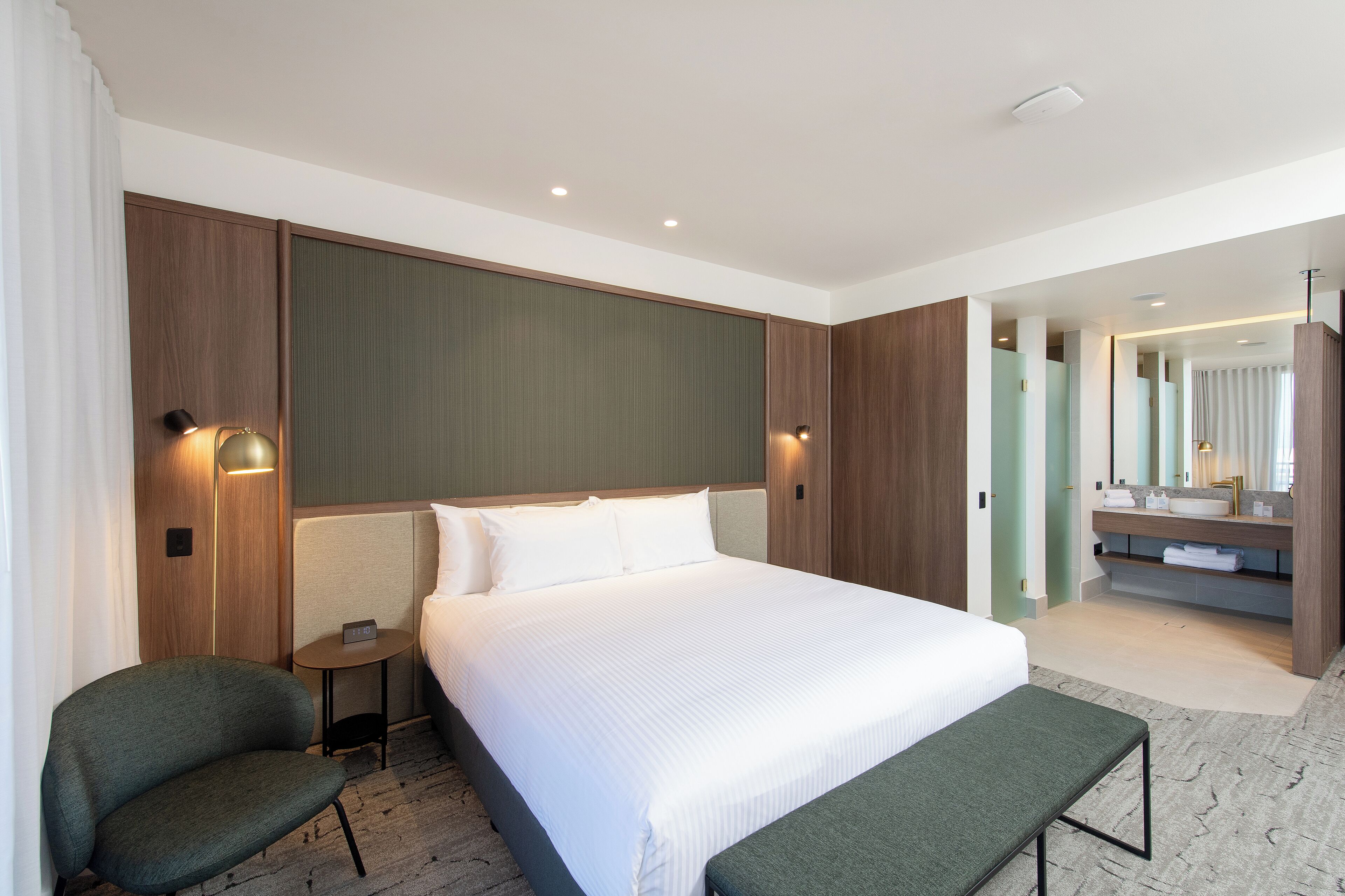 A modern hotel room with a large bed, a small bedside table, a chair, a bench, and a bathroom with a mirror and sink.
