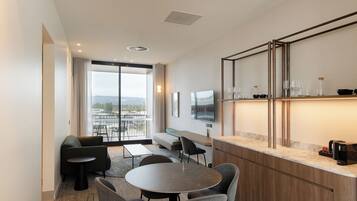 A modern hotel room with a dining area, a sofa, a TV, and a balcony with a city view.