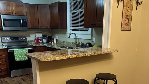 A kitchen with wooden cabinets, a granite countertop, a stainless steel oven, a microwave, and a sink. There are two black bar stools and a patterned rug on the floor. - Charming 2-bedroom apartment with wifi.   Three blocks to ferry.   (New Orleans)