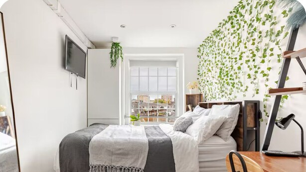 A bedroom with a bed, desk, chair, and a view of the city through a window.
