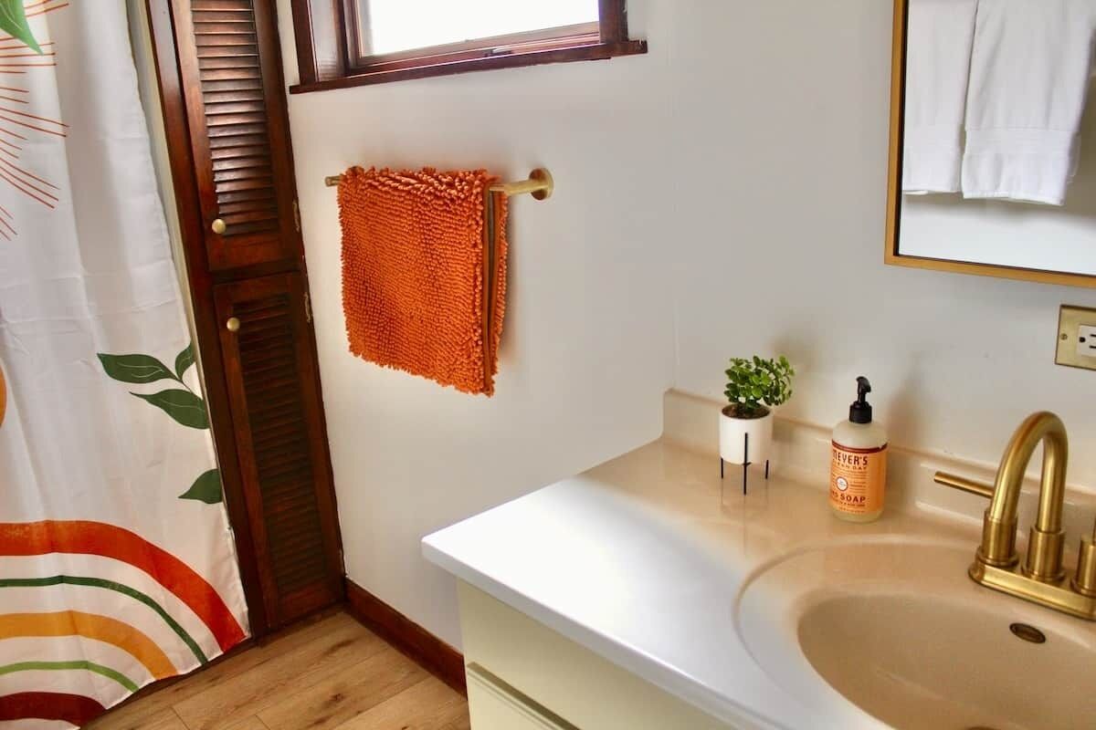 Un baño con un lavamanos blanco, un grifo dorado, un perchero con una toalla naranja, una pequeña planta en maceta y una botella de jabón.
