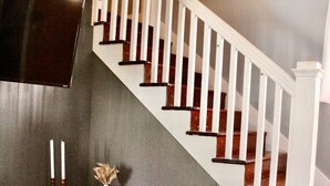 Eine Treppe mit weißen Geländern und Holzstufen, ein grauer Sessel mit einem gerollten orangefarbenen Kissen sowie ein dunkles Holzkonsole mit einer Vase und Kerzen.