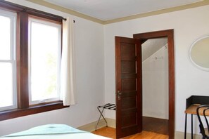 Una habitación con cama, ventana con cortinas, puerta que da a otra habitación y una pequeña mesa con silla.