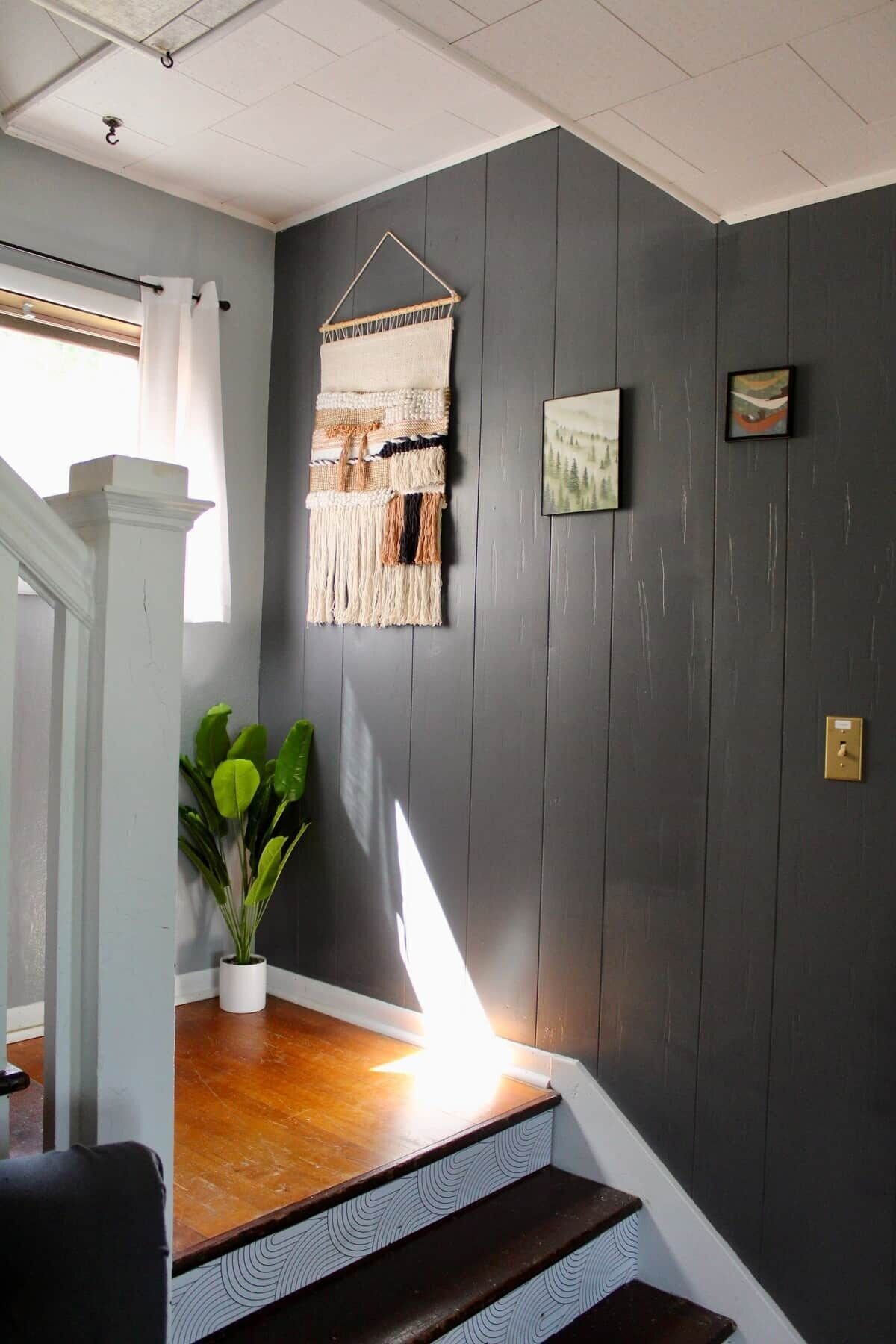 A staircase with a wooden finish, a wall-mounted macrame piece, a framed picture, and a potted plant.