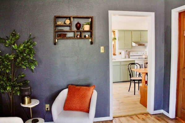 A room with a white armchair, a small side table, a wall-mounted shelf with decorative items, and a view into a kitchen area.