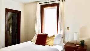 Ein Hotelzimmer mit Bett, Nachttisch mit Lampe, Fenster mit Vorhängen und Tür.