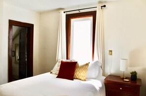 Una habitación de hotel con cama, mesita de noche con lámpara, ventana con cortinas y puerta.