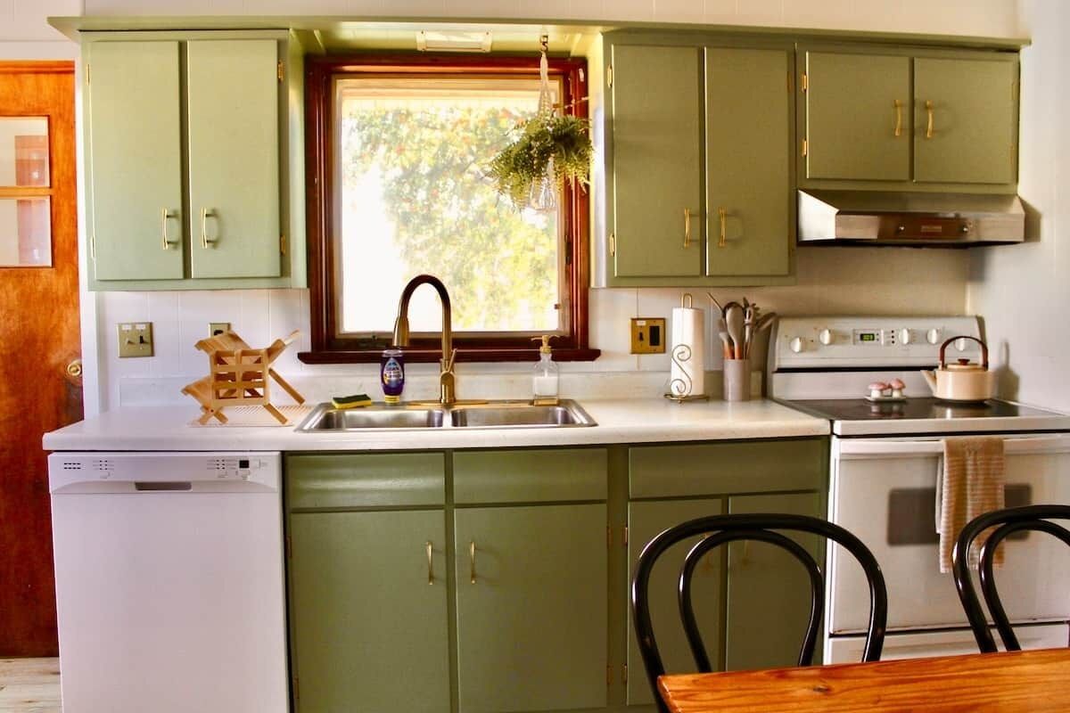 Una cocina con gabinetes verdes, un lavavajillas blanco, una estufa y una ventana con vistas a árboles.