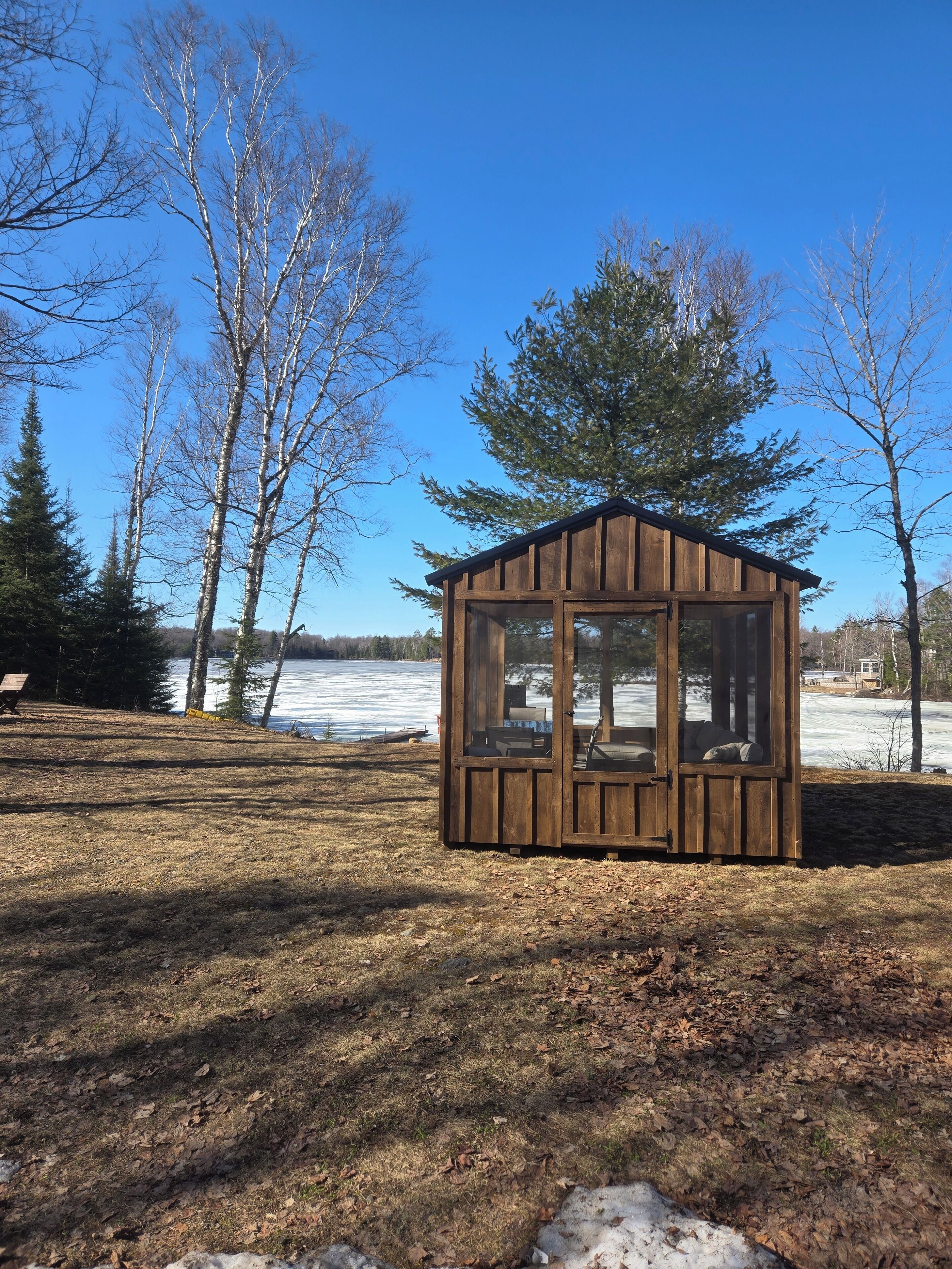 A small wooden cabin with a glass door and windows, surrounded by trees and a lake.