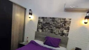 A bedroom with a bed, a headboard with a textured design, two wall-mounted lights, an air conditioning unit, and a nightstand with a phone and a bottle. - Sekhmatone 1-bedroom apartment (Luxor)