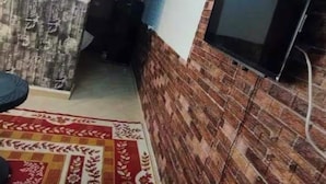 A narrow room with a patterned carpet, a television mounted on a brick wall, and a partially visible door. - Sekhmatone 1-bedroom apartment (Luxor)