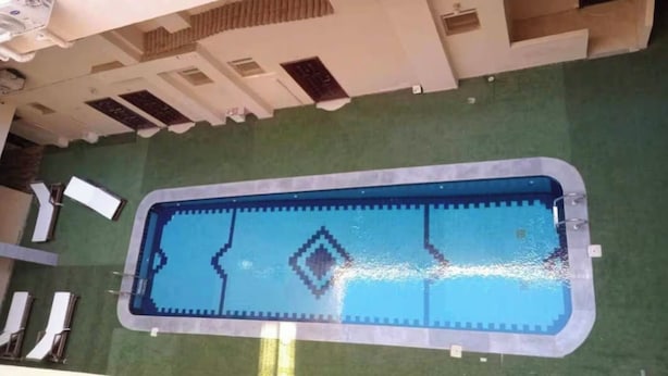 A rectangular swimming pool with a simple design, surrounded by a green lawn and a building with multiple windows and a flat roof. - Sekhmatone 1-bedroom apartment (Luxor)