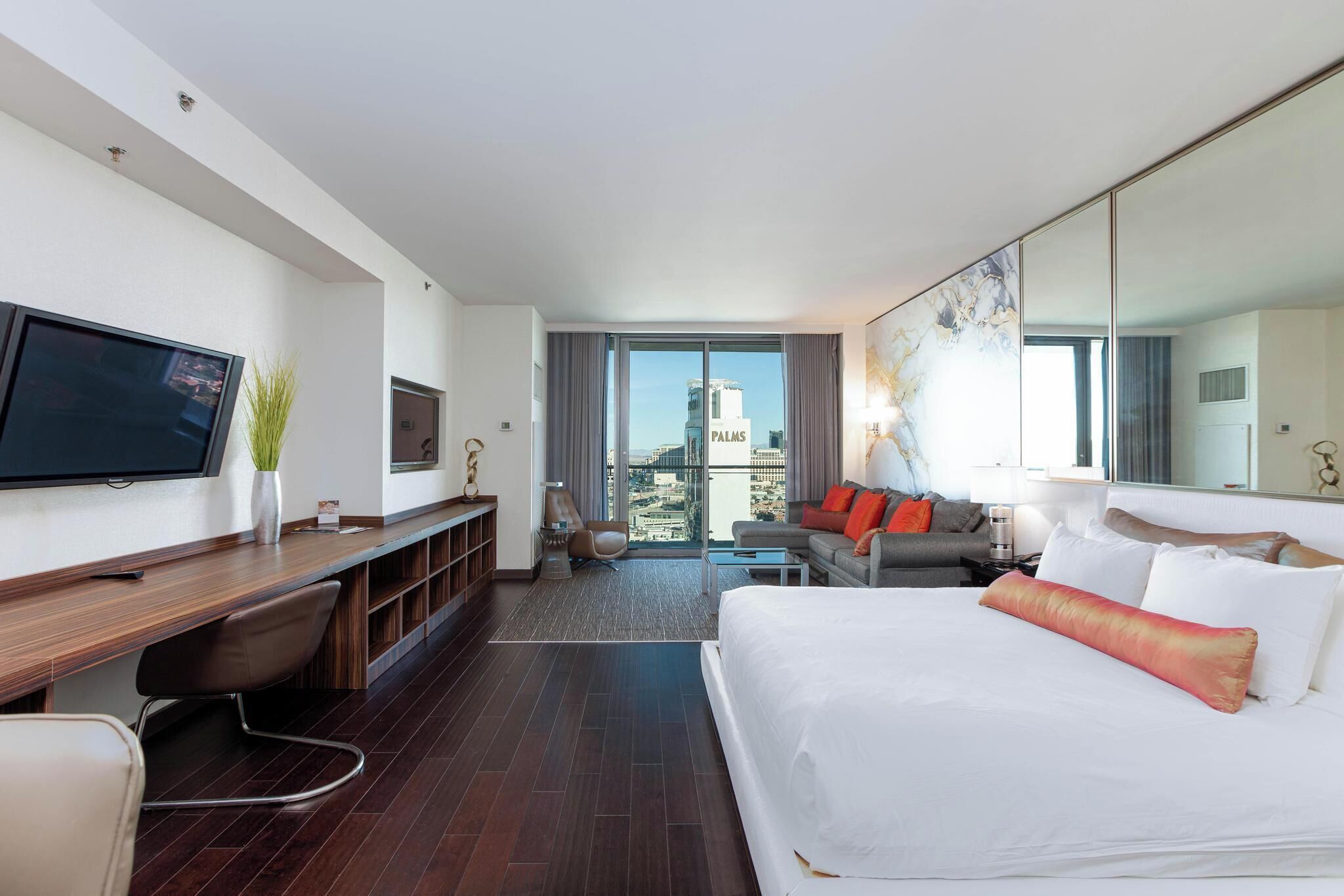 Une chambre d’hôtel moderne dotée d’un grand lit, d’un coin salon avec un canapé et des fauteuils, d’une télévision à écran plat, d’un bureau avec une chaise et d’une grande fenêtre offrant une vue sur la ville.