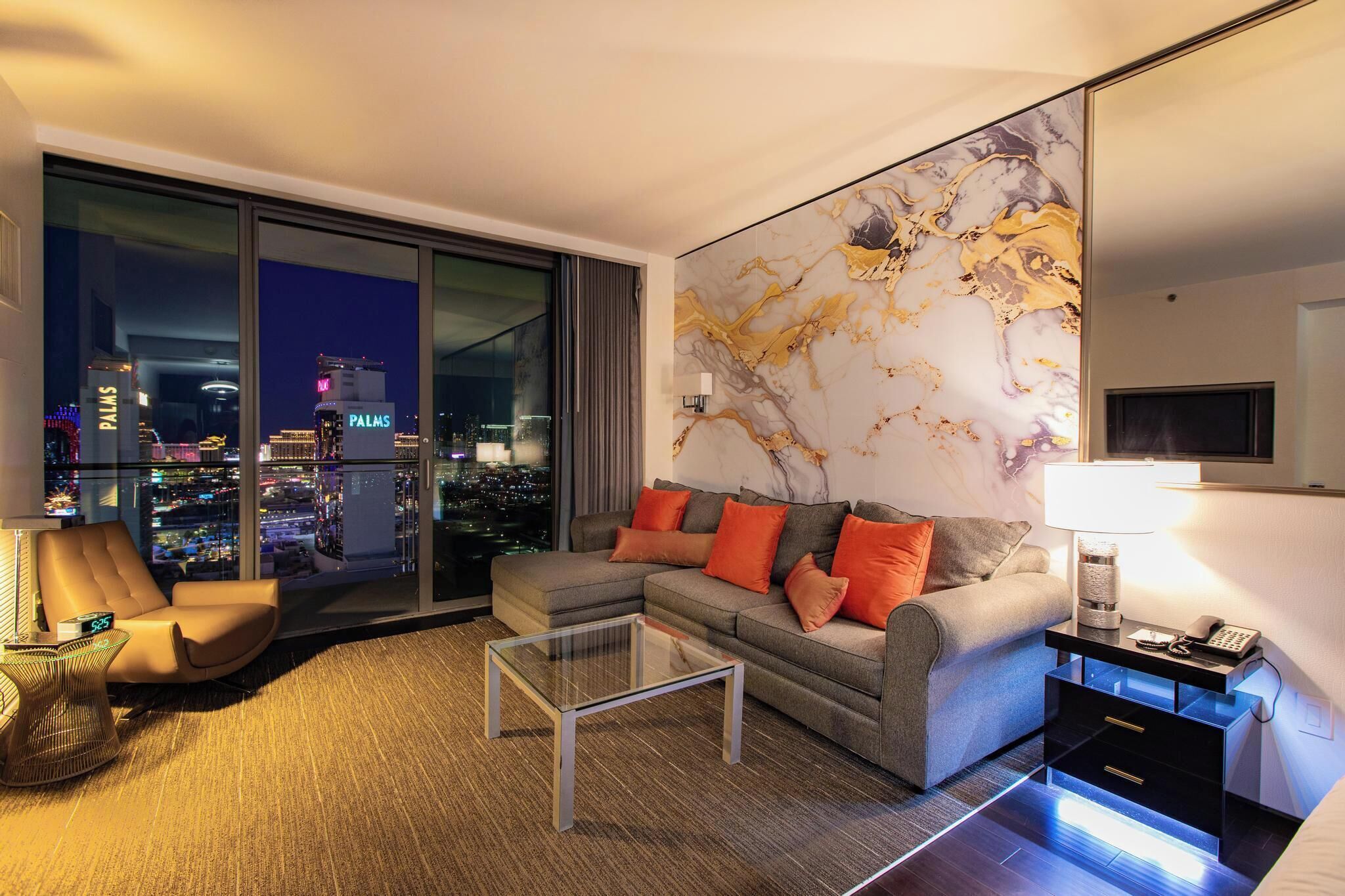 A modern living room with a city view, a sofa with orange cushions, a glass coffee table, and a nightstand with a lamp.