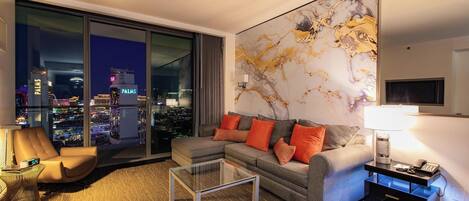 A modern living room with a city view, a sofa with orange cushions, a glass coffee table, and a nightstand with a lamp.