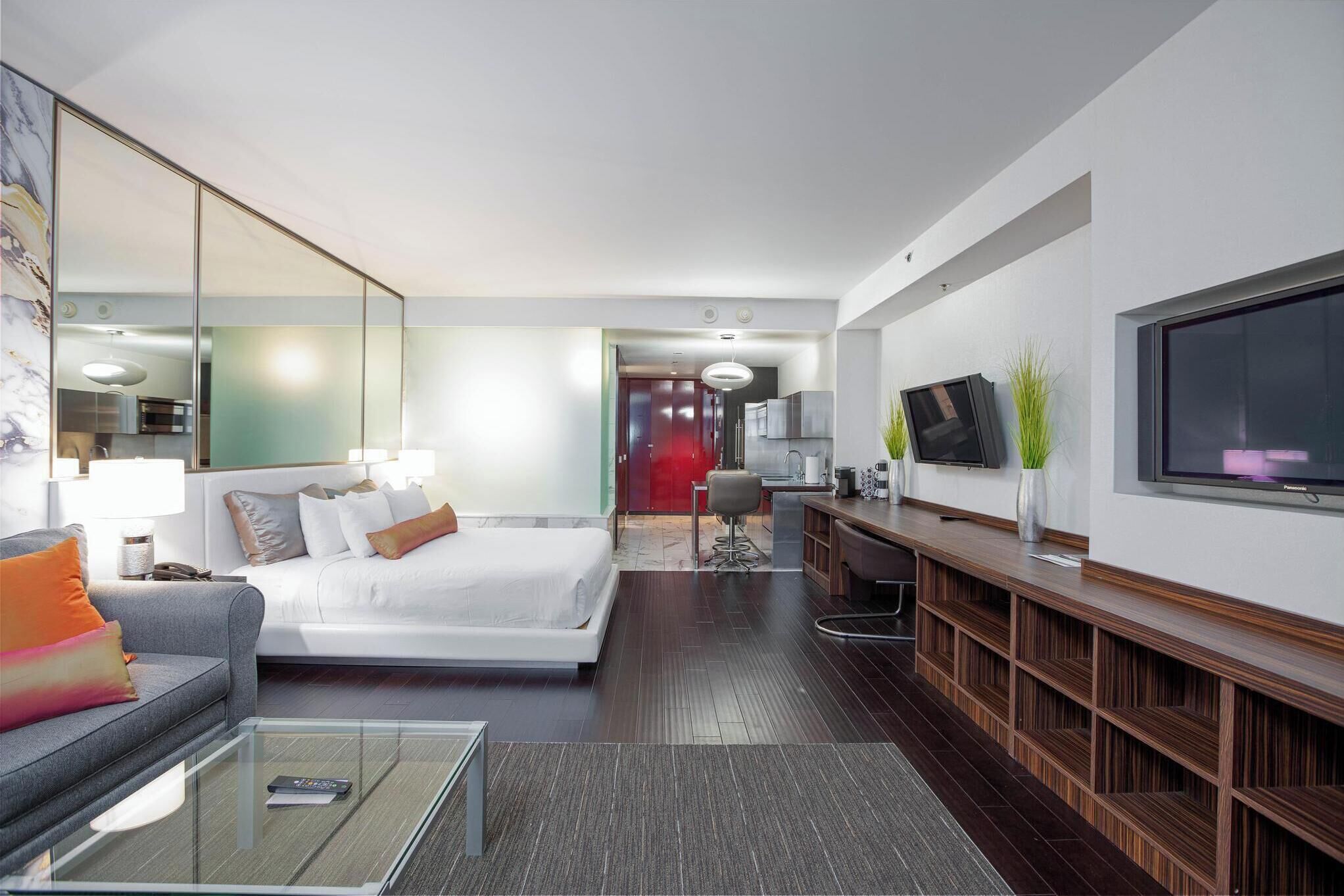 A modern hotel room with a large bed, a sofa, a TV, and a small kitchenette.