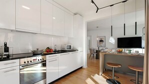 A modern kitchen with white cabinets, a black countertop, stainless steel appliances, and a wooden dining area with two bar stools.
