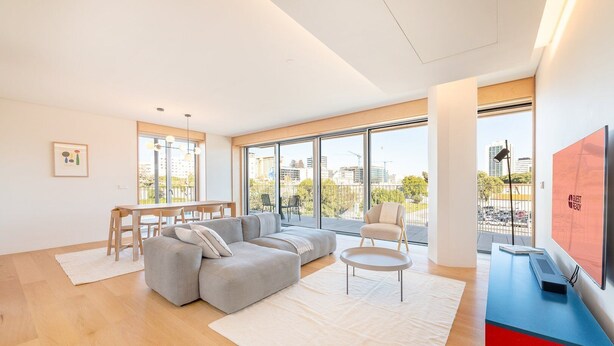 Una sala moderna con un sofá modular, una mesa de centro redonda y un comedor con mesa y sillas de madera. Amplias ventanas ofrecen vistas del paisaje urbano.