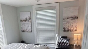 Une chambre à coucher comprenant un lit, une table de chevet, une lampe, une fenêtre avec des stores et des œuvres d’art accrochées aux murs.