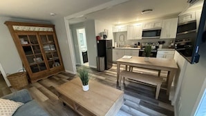 A modern kitchen with stainless steel appliances, a dining area with a wooden table and bench, and a living room with a sofa and a coffee table. - Driftwood Getaway - Perfect Location Near Beach! (Hampton)