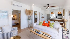 A modern living room with a white sofa, striped armchair, and a wooden coffee table. There is a kitchen area with white cabinets and a dining table. A ceiling fan is visible, and there are decorative items like a vase with dried plants and a framed picture on the wall.