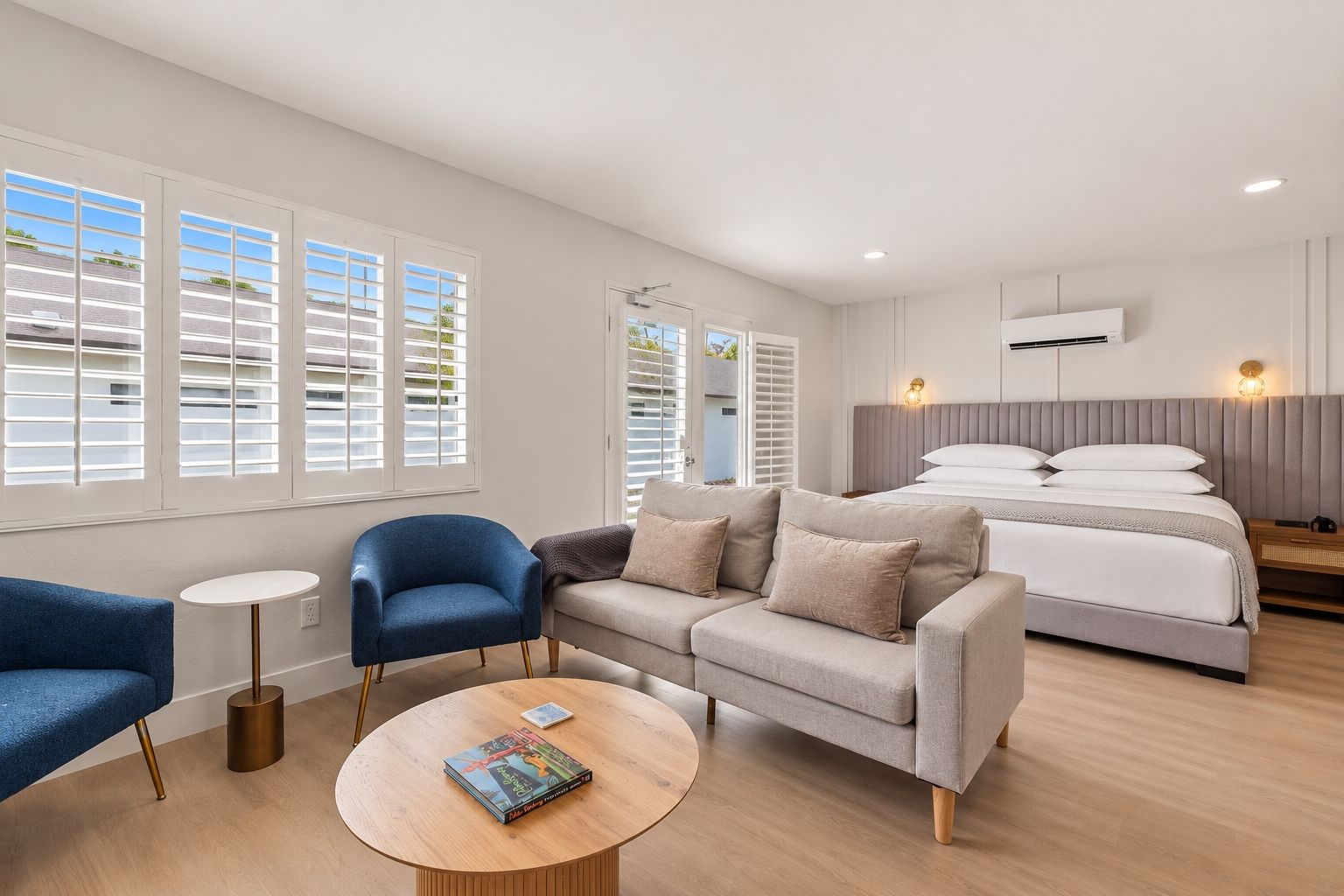 Een moderne hotelkamer met een bed, twee sofa's, een ronde salontafel en grote ramen met zonwering.