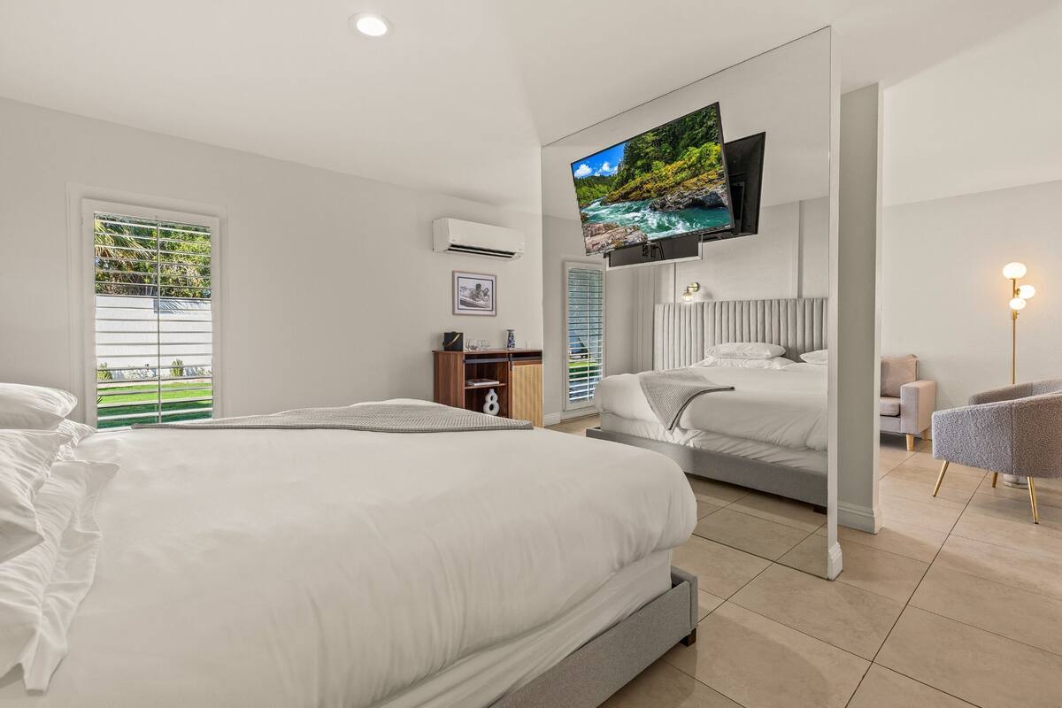 A modern hotel room with a large bed, a TV mounted on the wall, and a view of greenery outside.