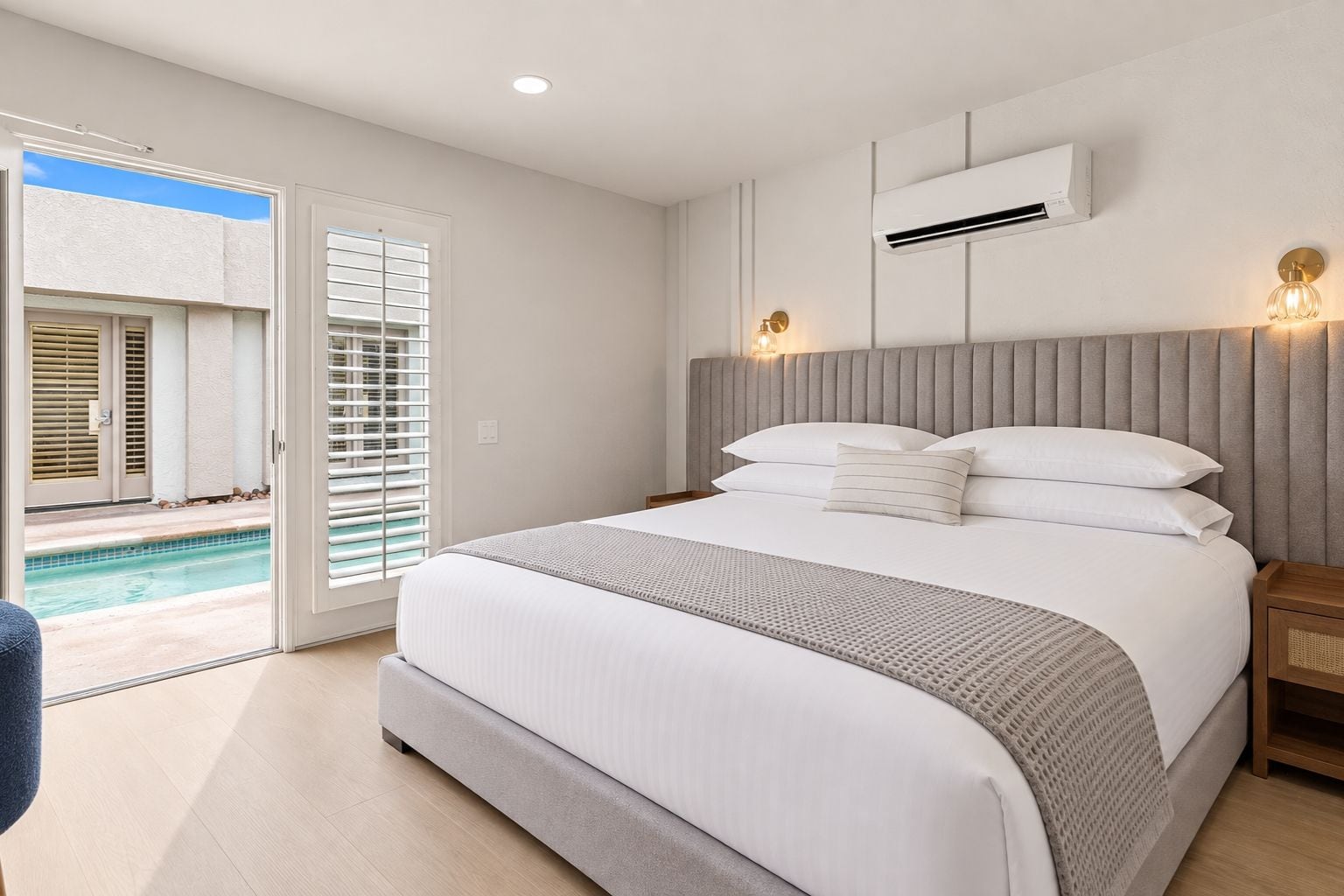 Quarto de hotel com uma cama grande, mesa de cabeceira, aparelho de ar condicionado e vista para uma piscina através de uma porta de vidro deslizante.