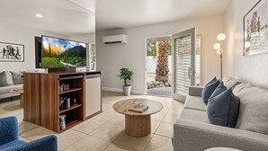 Una sala moderna con un televisor de pantalla plana, un mueble de entretenimiento de madera, una mesa de centro redonda, un sofá gris y un ventanal con vista a un jardín.
