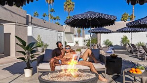 Una pareja relajándose en un sofá cerca de un brasero, con asientos exteriores y sombrillas en un espacio al aire libre soleado.