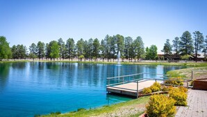 Un lago tranquilo con un muelle de madera y una fuente, rodeado de árboles y un cielo despejado.