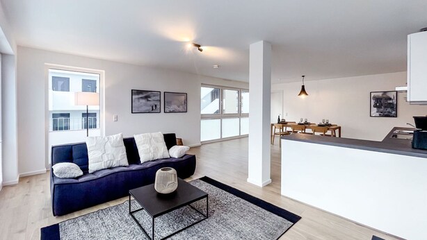 A modern living room with a dark blue sofa, a black coffee table, and a kitchen area with a white countertop and wooden chairs. - Vacation apartment in the mountain resort 3.3.5 - Adventure & wellness with infrared cabin! (Winterberg)