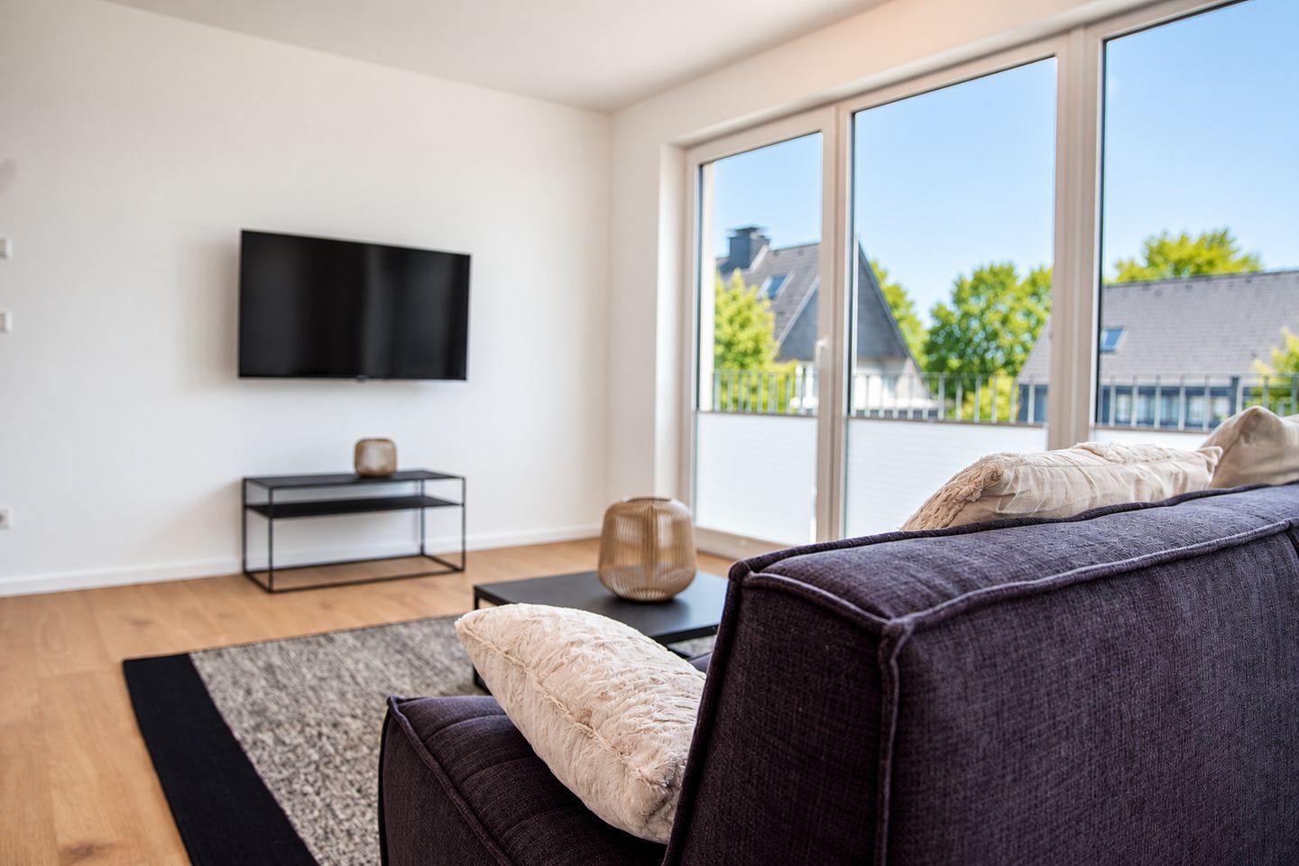 A modern living room with a large window, a flat-screen TV mounted on the wall, a dark sofa, and a small side table with a vase.