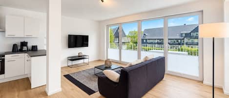 Un espacio habitable moderno con cocina, televisor de pantalla plana, sofá y ventanas amplias que ofrecen vista a una zona residencial.