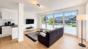 A modern living space with a kitchen, a flat-screen TV, a sofa, and large windows offering a view of a residential area. - Vacation Apartment in a Mountain Resort 3.3.5 - Adventure & Wellness with an Infrared Sauna! (Winterberg)