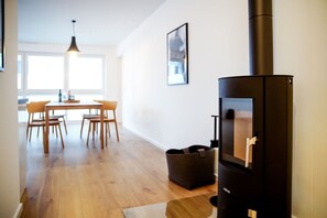 Un salon moderne doté d’un poêle à bois, d’un coin repas avec des chaises et une table en bois, et d’une grande fenêtre laissant entrer la lumière naturelle.