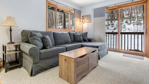 Una sala moderna con un sofá modular, una mesa de centro de madera, una lámpara de pie y ventanas amplias con vistas a árboles.