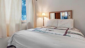 Un dormitorio con cama, lámpara de noche, ventana con cortinas y espejo en la pared.