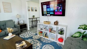 Una sala de estar con un televisor de pantalla plana grande montado en la pared, un sofá gris, una mesa de centro marrón con un control remoto, un tapete estampado y un mueble blanco con estantes.