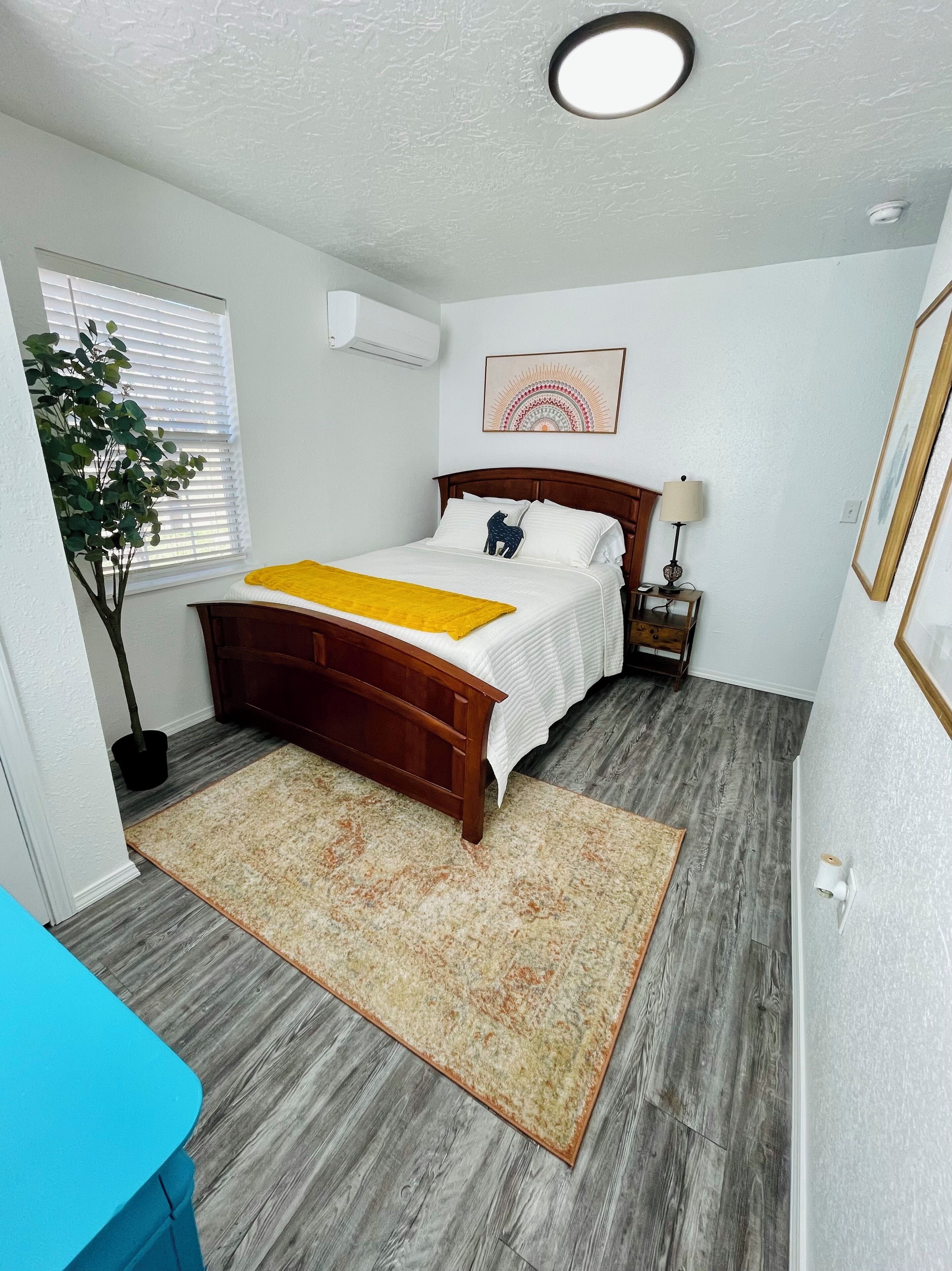 Un dormitorio con una cama de madera, una mesita de noche con lámpara, un aire acondicionado de pared, una ventana con persianas y una planta en maceta.